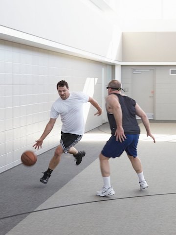 Two men play basketball indoors. One dribbles the ball across the three point line while the other tries to defend.
