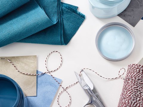 An overhead view of blue and tan fabric swatches stacked on top of each other by a folded blue fabric sample, a red and white thread, and scissors.