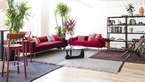 Um sofá e um sofá vermelho Bolster ancoram um ambiente residencial que inclui bancos, plantas e estantes de livros.