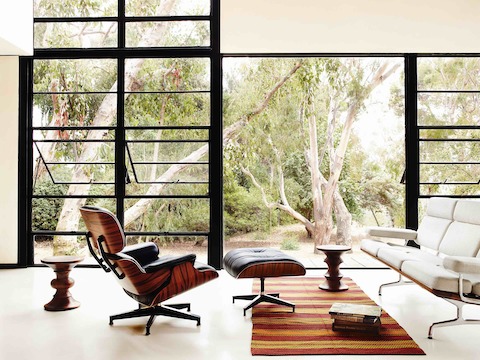 A black leather Eames Lounge Chair and Ottoman, Eames Sofa, and two Eames Walnut Stools near a glass wall. 