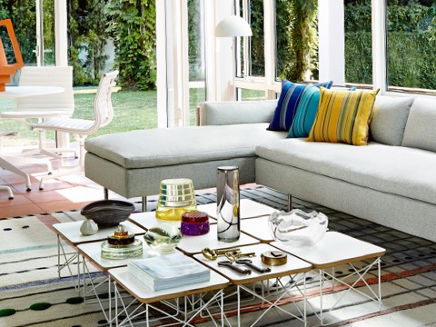 A residential setting with floor-to-ceiling windows, a gray Bolster Sofa, and nine Eames Wire Base Low Tables with white tops. 