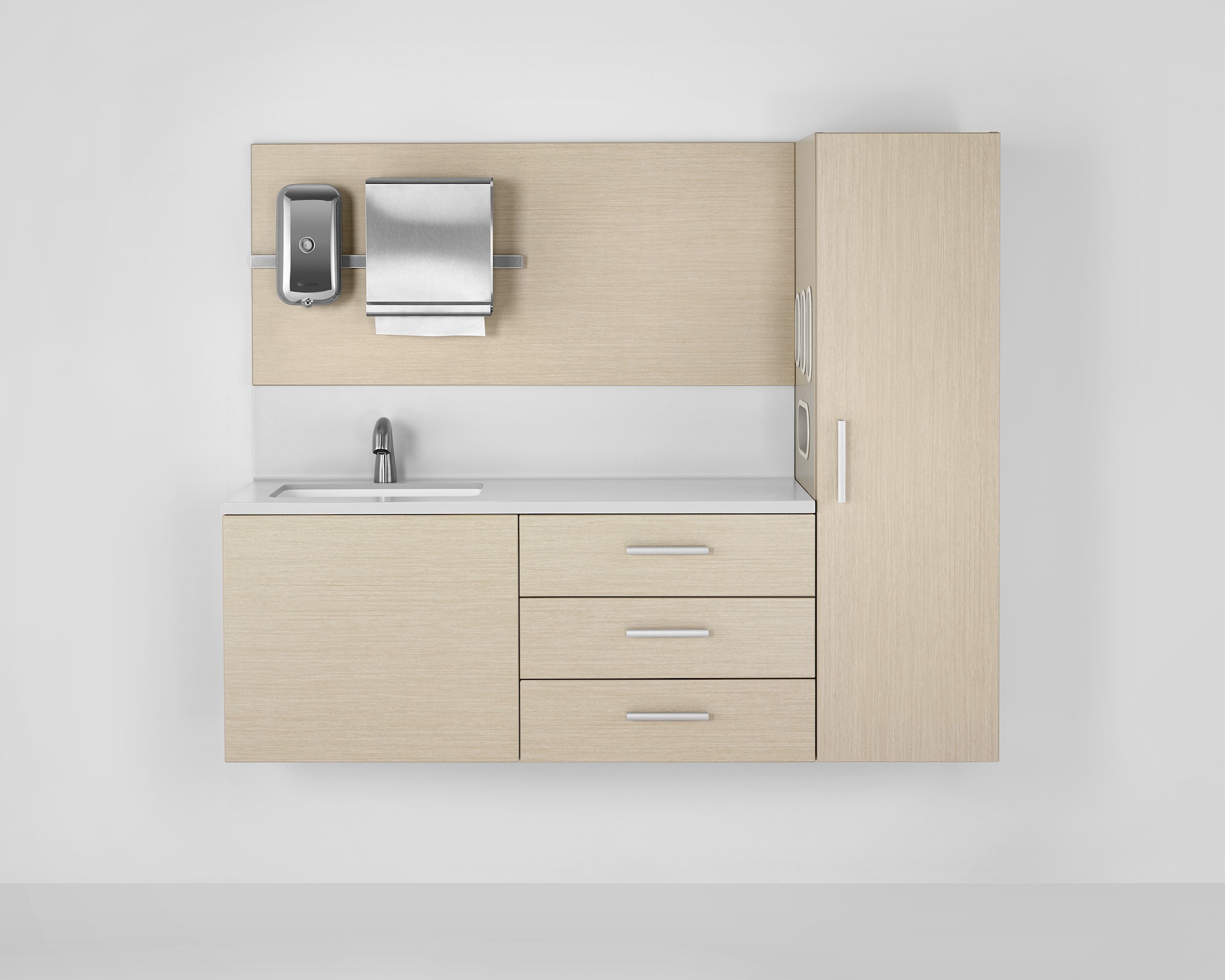 A Mora casework wall in an ash finish consisting of a 3-drawer storage case, a waredrobe, and a wall panel with a soap dispenser and paper towel dispenser. The white solid surface has an integrated sink and backsplash.