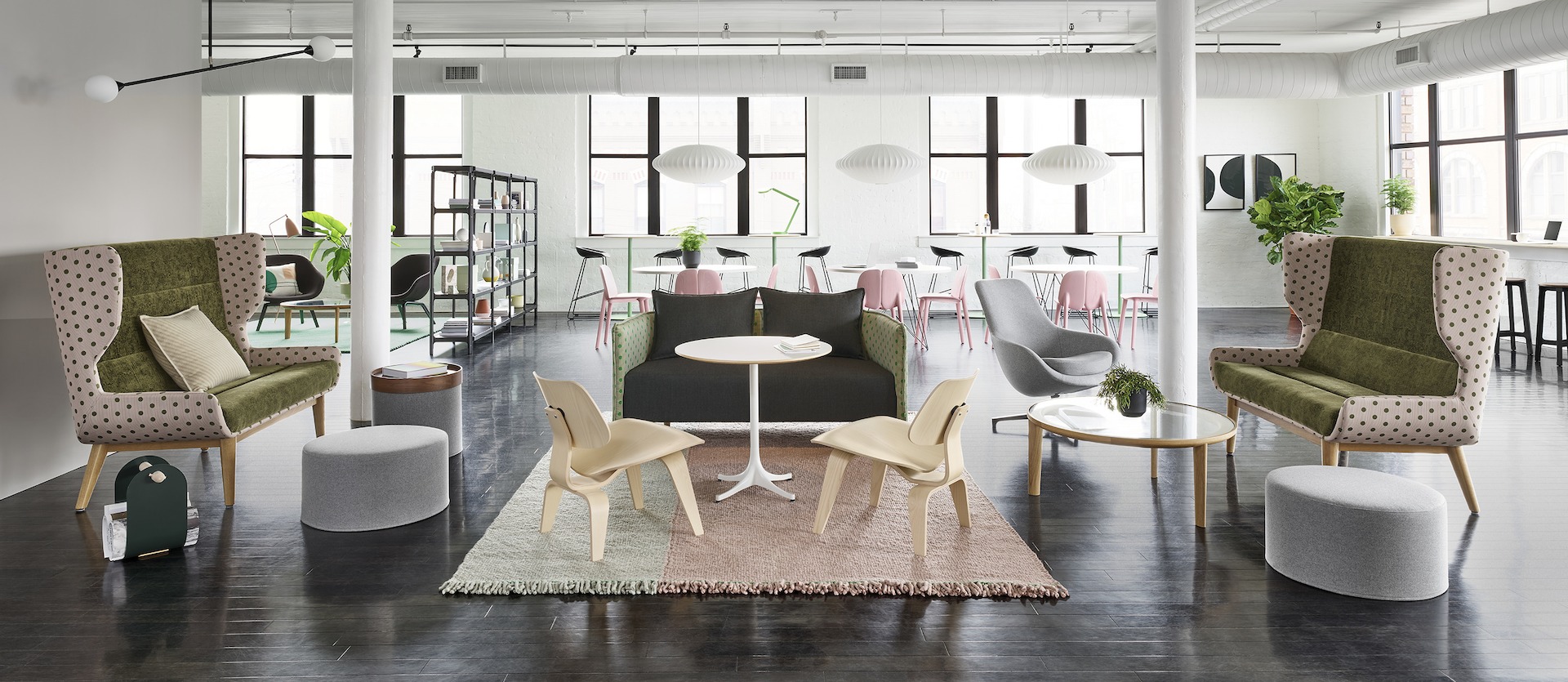 Two Hush Two Seat Sofas with wood legs in pink and green textiles frame a lounge space. In the center of the space is a Hush Two Seat Sofa in dark grey and avocado Maharam Dot textiles. Additionally in the space are two Eames Molded Plywood lounge chairs, a white Nelson Pedestal Table, a Port Collection Coffee Table with an oak base and a glass top, three light grey Drum Poufs, and a salmon and tan rug. The scene is set in front of various collaborative areas and large windows. 