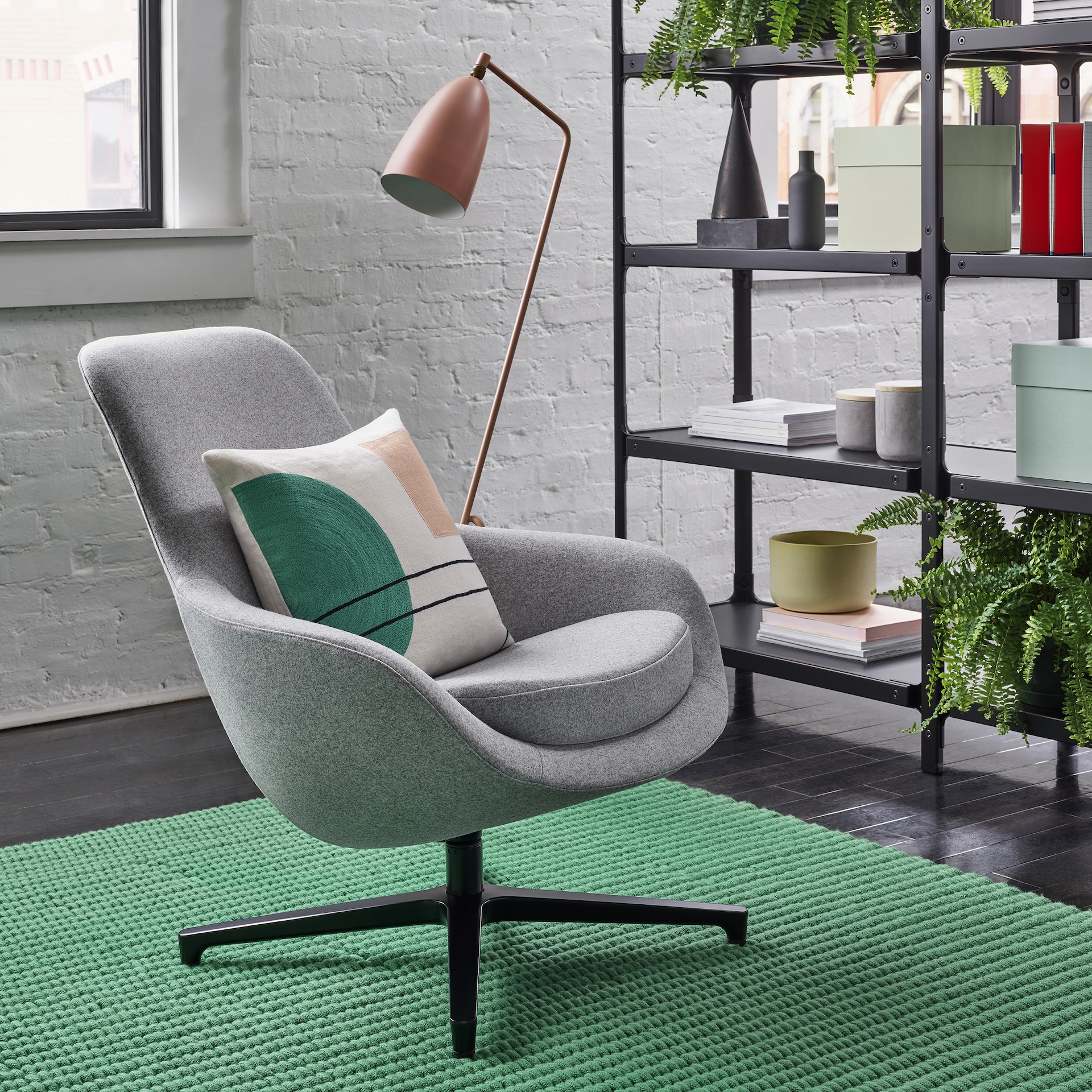 A light grey Saiba Lounge Chair with a black base on a green rug in front of  a black Steelwood Shelving System and a salmon Grasshopper Floor Lamp. The scene is set in front of a white wall with windows. 