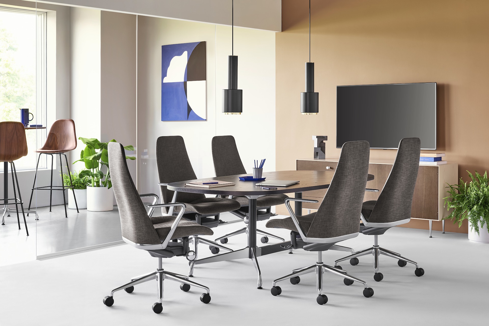 A meeting space setting featuring four black Taper Chairs surrounding an ebony Eames Table. A Nelson Thin Edge Chest sits beneath a TV against the back wall and two Eames Molded Wood Stools cluster around a tall table in the background. 