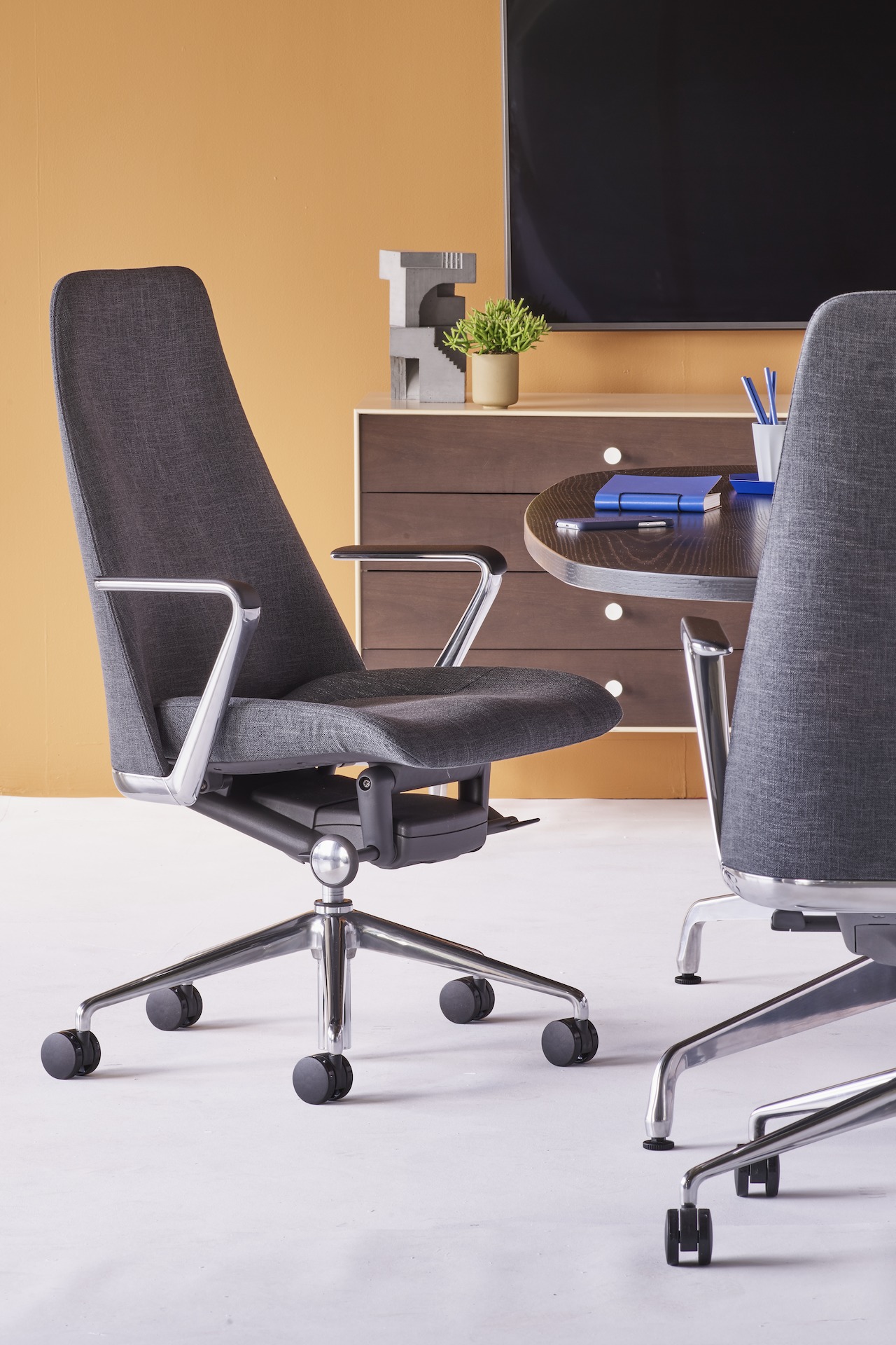 A meeting space setting featuring two black Taper Chairs surrounding an ebony Eames Table. A Nelson Thin Edge Chest sits beneath a TV against the back wall.