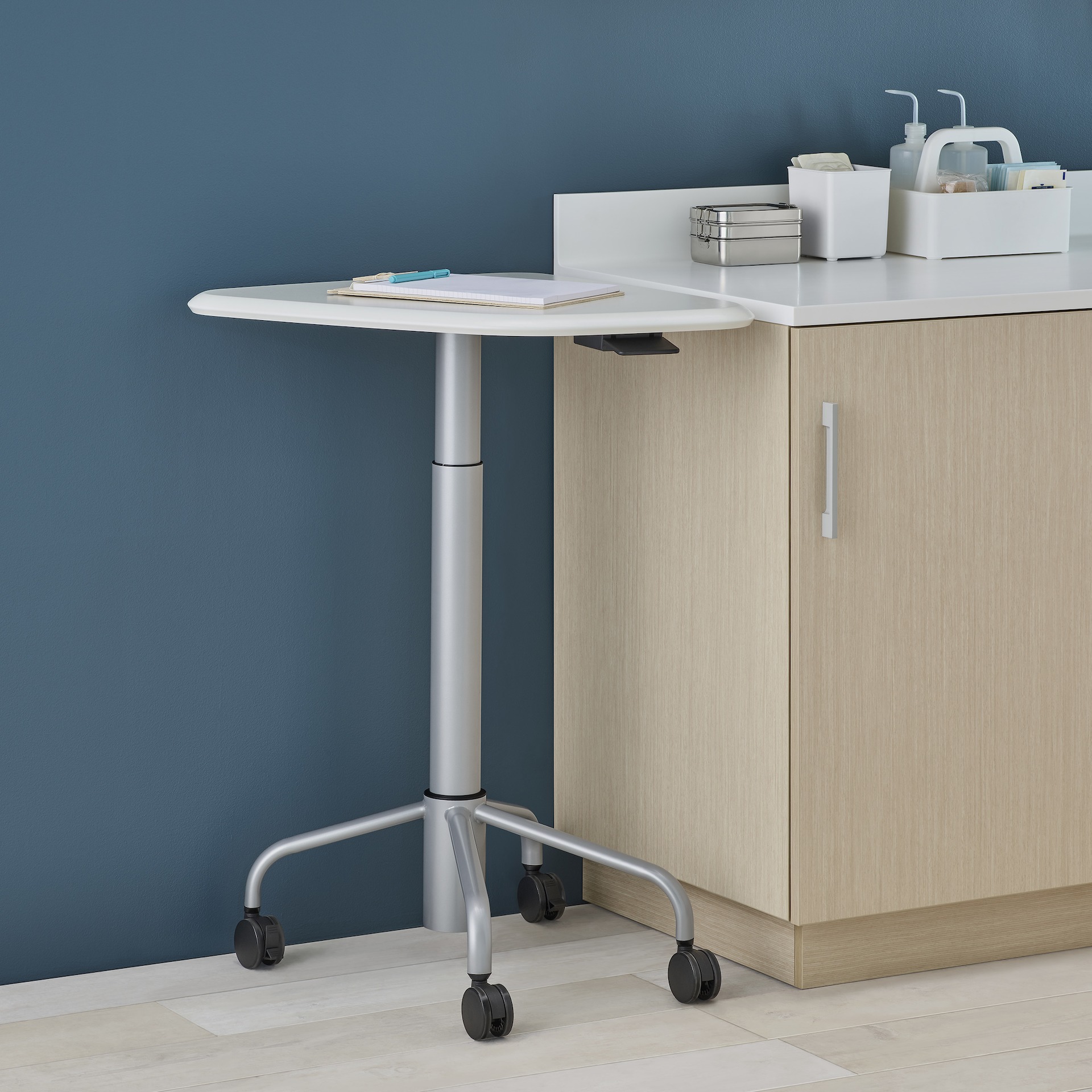 An Intent Solution height-adjustable mobile technology table with a white surface and silver base next to Mora System casework in a light wood finish and Corian surface.