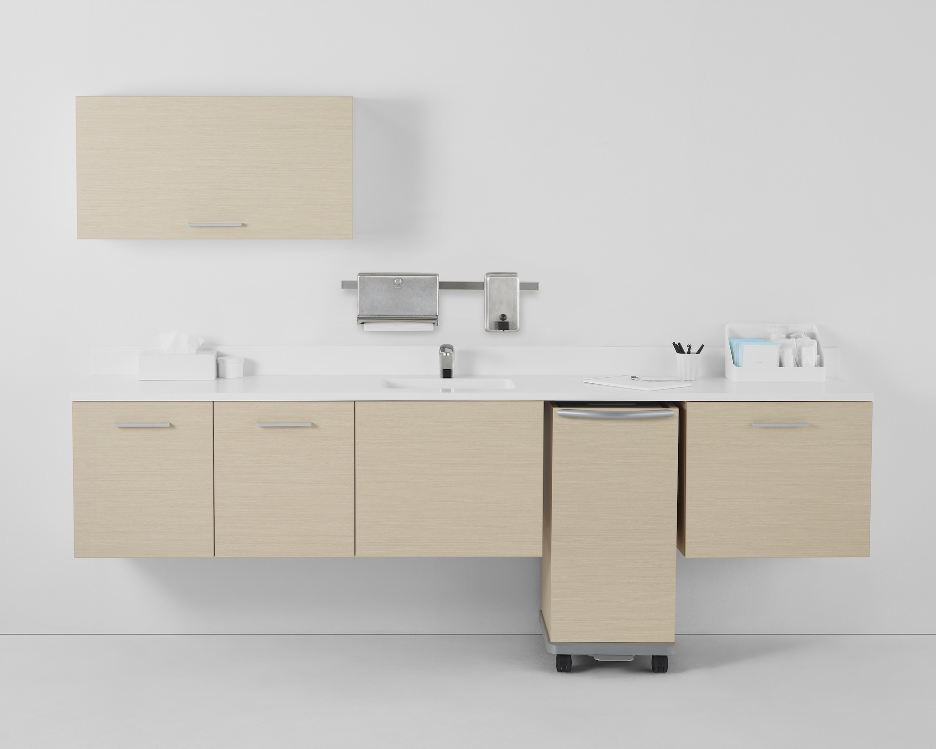 A Mora casework wall in a light ash finish consisting of one overhead storage cabinet and three lower supply cabinets with a linen cart docked within the casework.