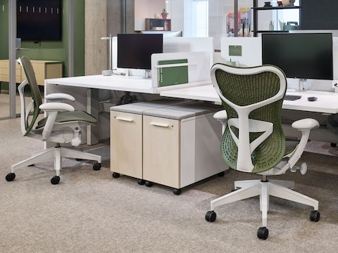 Two olive green and white work chairs sit at a white and light wood workstation with computer monitors and desktop accessories, and rolling storage underneath.