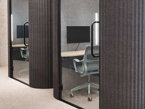 Two dark gray rounded work pods with glass doors and sleek black handles sit in an open office, both with light wood work surfaces, a computer monitor, and light gray work chairs inside.