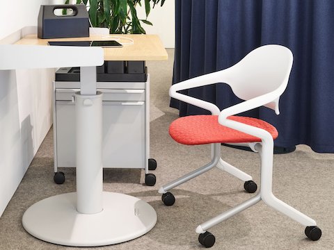 A red and white rolling office chair sits at a square wood and white sit-to-stand table with dark gray powerbox and accessories atop it it, with large plant and dark blue curtains in the background.