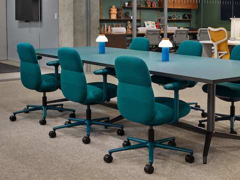 Six teal upholstered work chairs sit at a long teal conference table with two small blue lanterns on the surface, in an open office setting.