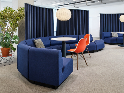 A dark blue modular sofa in a curving shape sits in front of dark blue hanging curtains. Two small round tables with red shell chairs accompany the sofa, and bubble pendants hang above, with large plants behind.