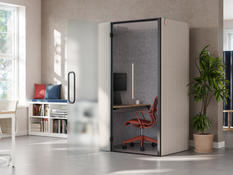 An open office, featuring an enclosed Bay Work Pod with red task chair pulled up to a work surface with computer monitor.