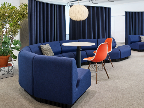 A dark blue modular sofa in a curving shape sits in front of dark blue hanging curtains. Two small round tables with red shell chairs accompany the sofa, and bubble pendants hang above, with large plants behind.