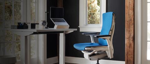 Home Office com uma Embody Chair azul e uma mesa ajustável Sit-to-Stand Desk, destacando o texto 'Outlet de verão' com descontos de até 50% em designs selecionados. 