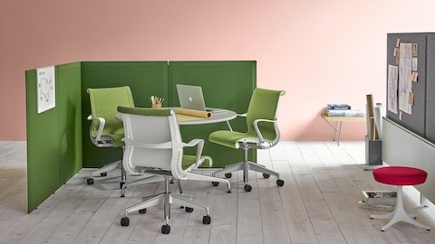 Green screens define a partially enclosed meeting area that contains a round table and green Setu office chairs.