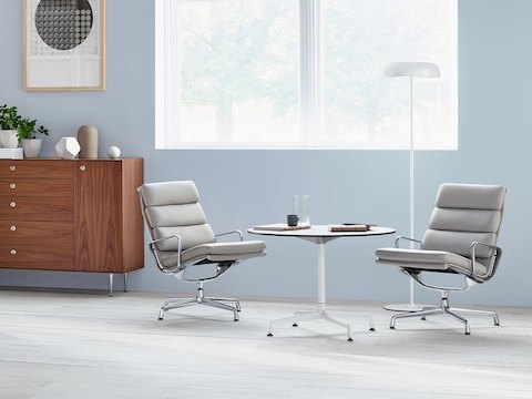 2つの明るい灰色のEames Soft Padラウンジチェアと白いフレームと上部の丸いEamesテーブル。