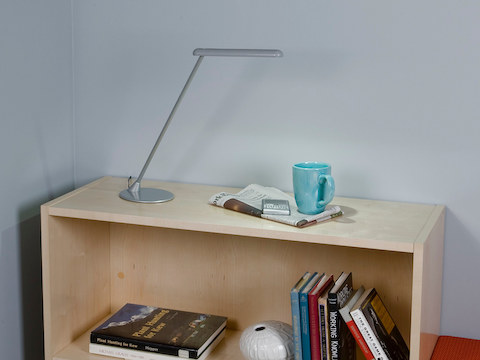 A silver freestanding Flute Personal Light, newspaper, and coffee cup atop a low bookcase.