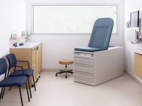 Mora casework in an oak finish with a mobile supply cart and a mobile trash cart with the caregiver work area, a technology support cabinet, two Verus Side Chairs in blue, a physician stool in tan, and an exam table.