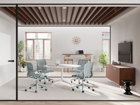 Four glacier blue Setu Chairs surround a circular table in front of a monitor in a glass-enclosed meeting room.