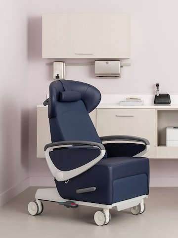An exam room with an Ava Recliner in a dark blue upholstery and white base and arms with urethane arm caps and Mora System casewok behind it in a light ash finish.
