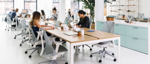A handful of colleagues work independently in an open office that features a benching setup with light grey Sayl office chairs.