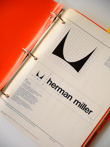 A red three-ring binder open to a page with a large Herman Miller logo in black and information about brand standards.