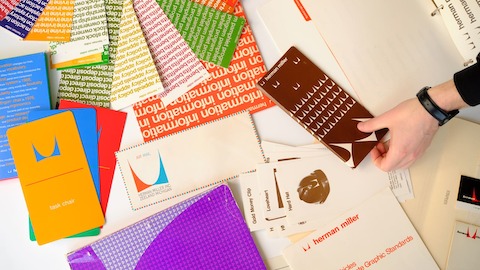 Colorful brochures from Herman Miller's archive are displayed loosely on a white tabletop with a hand picking up one.
