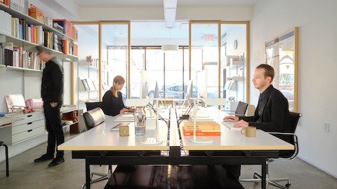 Three people dressed in black work around a large white conference table with black office chairs, with neatly displayed books on wall shelving in front of a large window looking out to a street view.