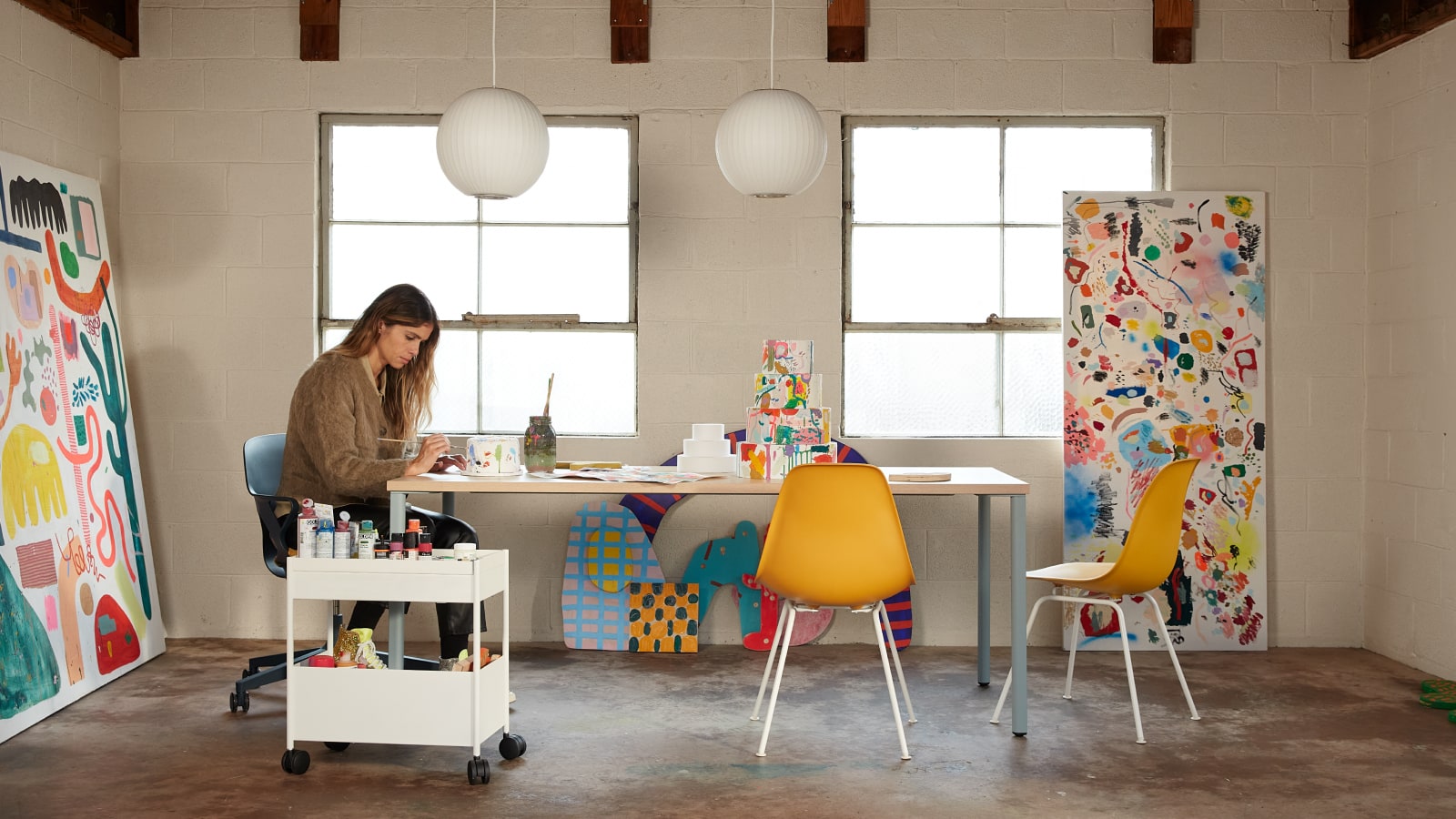 Artist Kindah Khalidy sitting in a Zeph Chair at a table, with two yellow Eames shell chairs.