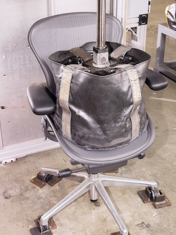 A dark gray work chair sits in a cement testing facility, with a large weighted black bag sitting in its seat.