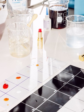 Glass beakers with different colored liquids, a red lipstick tube, glue bottle, dish soap bottle sit on a tabletop with numbered squares ready for testing.