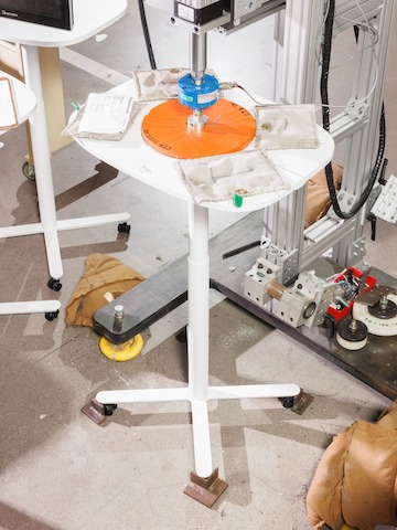 A small white height-adjustable table is weighted down with sand bags and a circular orange weight on a metal arm in a testing facility.