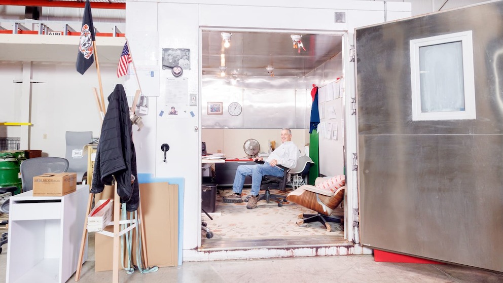 A large white chamber sits inside the Test Lab warehouse, with its aluminum door opened wide. Inside is a makeshift office space, where a man sits in his office chair at a desk.