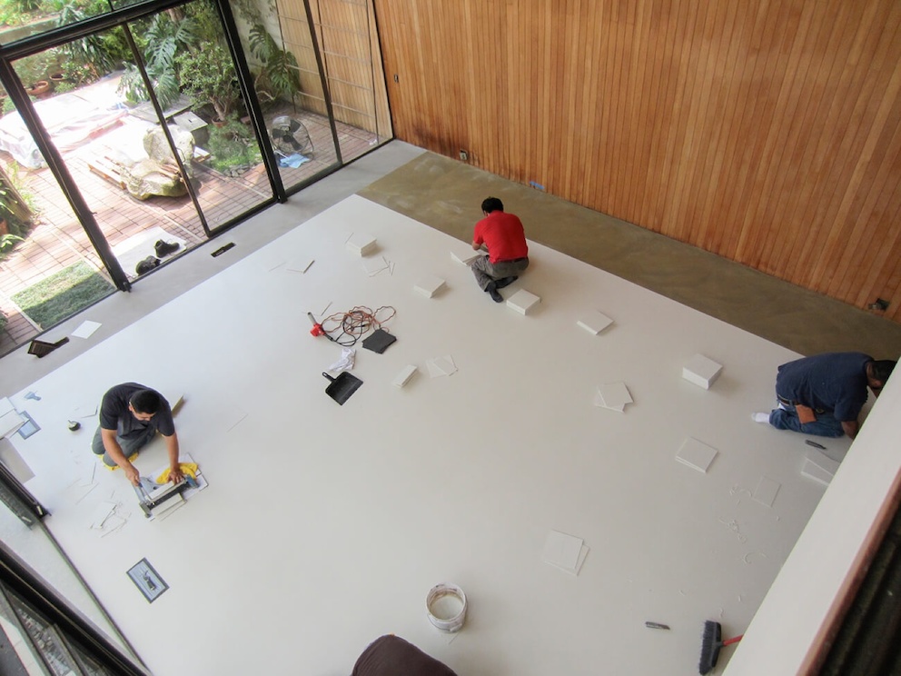 A color photo of a bird's eye view of three men sitting on a covered floor in the Eames House living room. They use tools to work on repairs to the space.